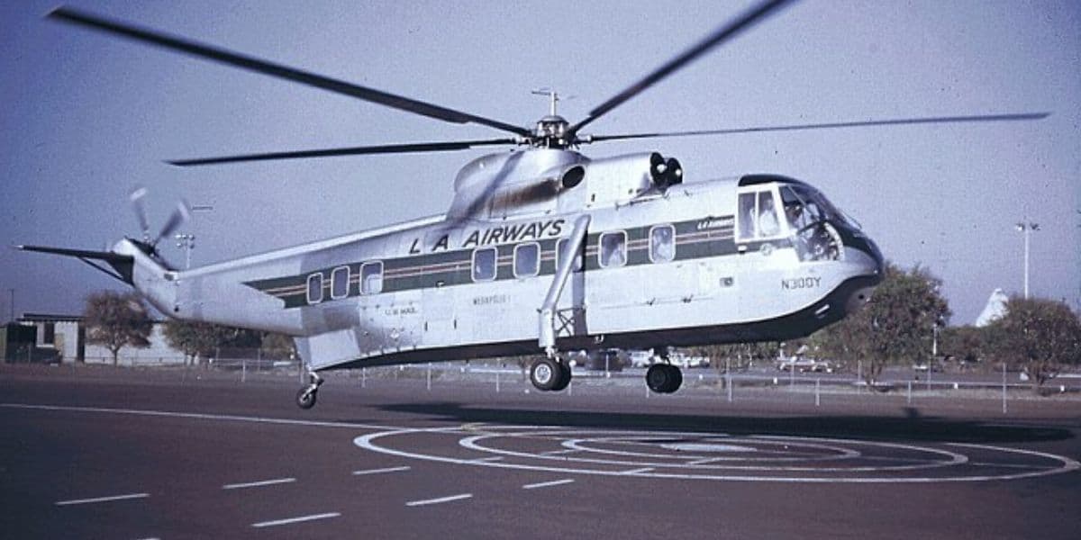 A vintage helicopter bearing L.A. Airways is hovering above a designated landing area. The gray helicopter has a stripe, set against a clear blue sky.